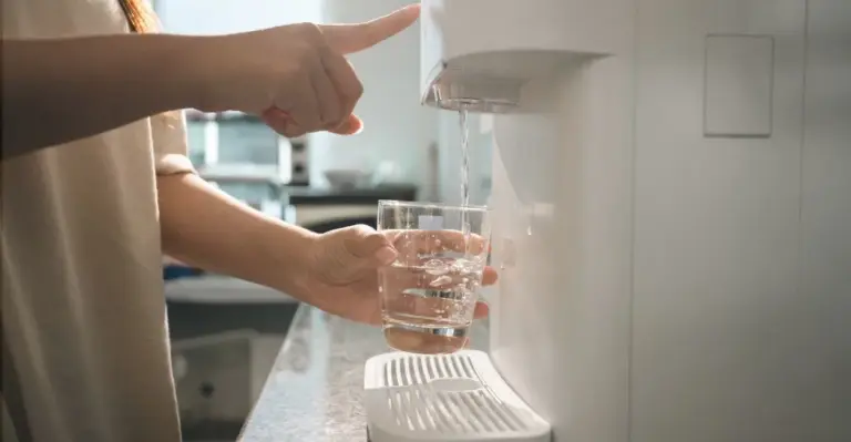 Pessoa enchendo um copo de água em um purificador de água moderno, visto de lado, mostrando apenas os braços, o copo transparente e o filtro branco. quais os melhores purificadores de agua